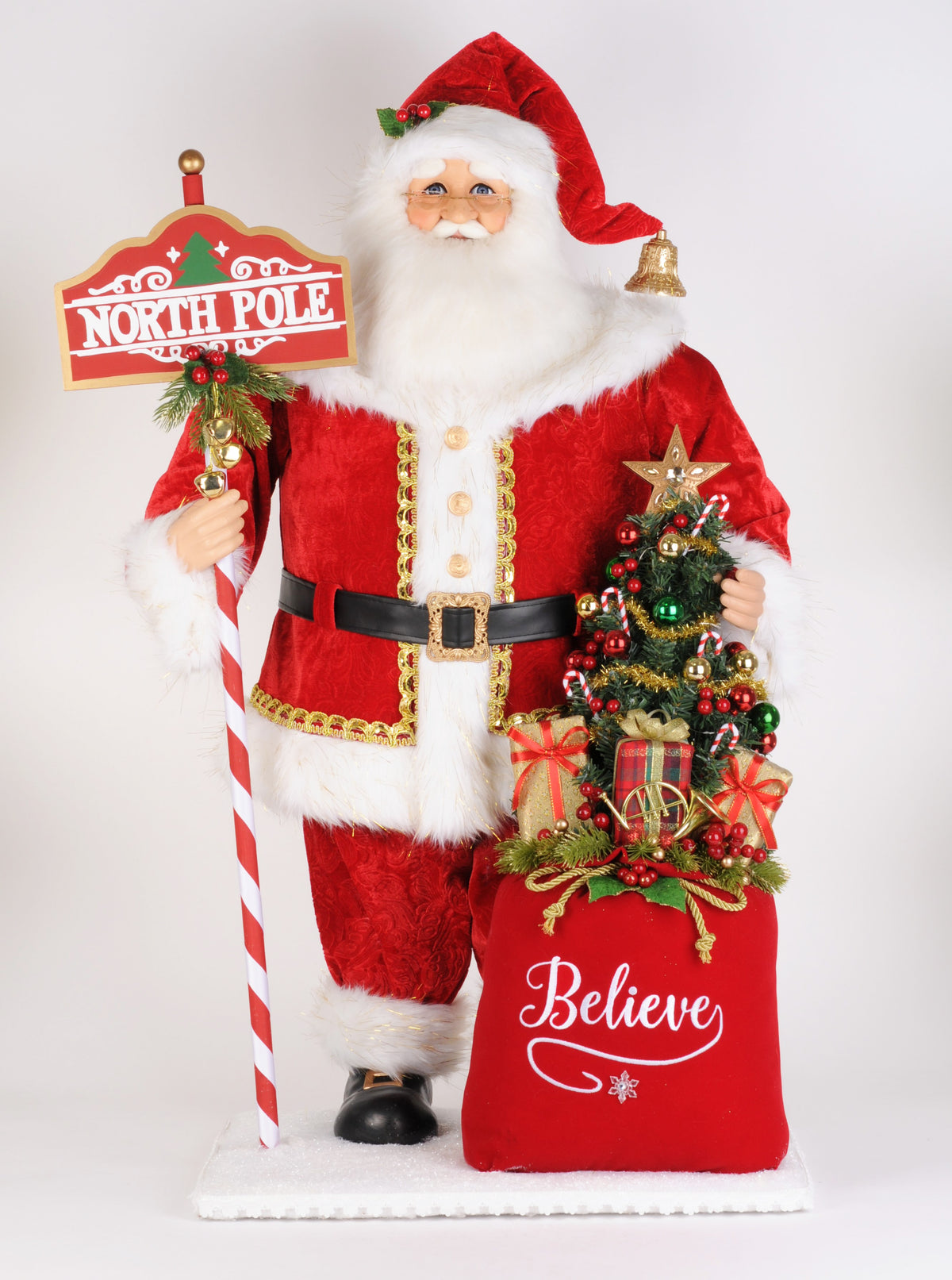 Santa figurine in a red suit with white fur trim, holding a North Pole sign beside a Believe gift sack.