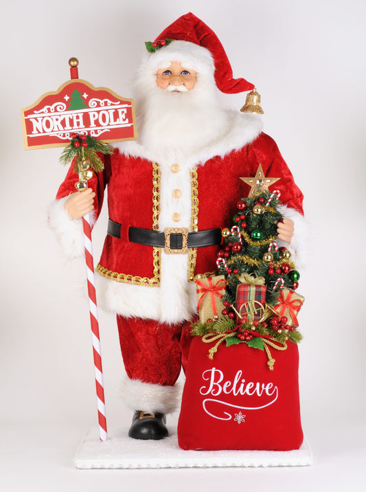 Santa figurine in a red suit with white fur trim, holding a North Pole sign beside a Believe gift sack.